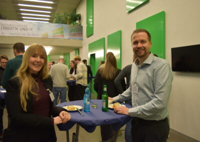 blonde Frau und Mann mit blonden Haaren stehen an Stehtisch bei einem Gala-Buffet, im HIntergrund Menschen, die sich unterhalten