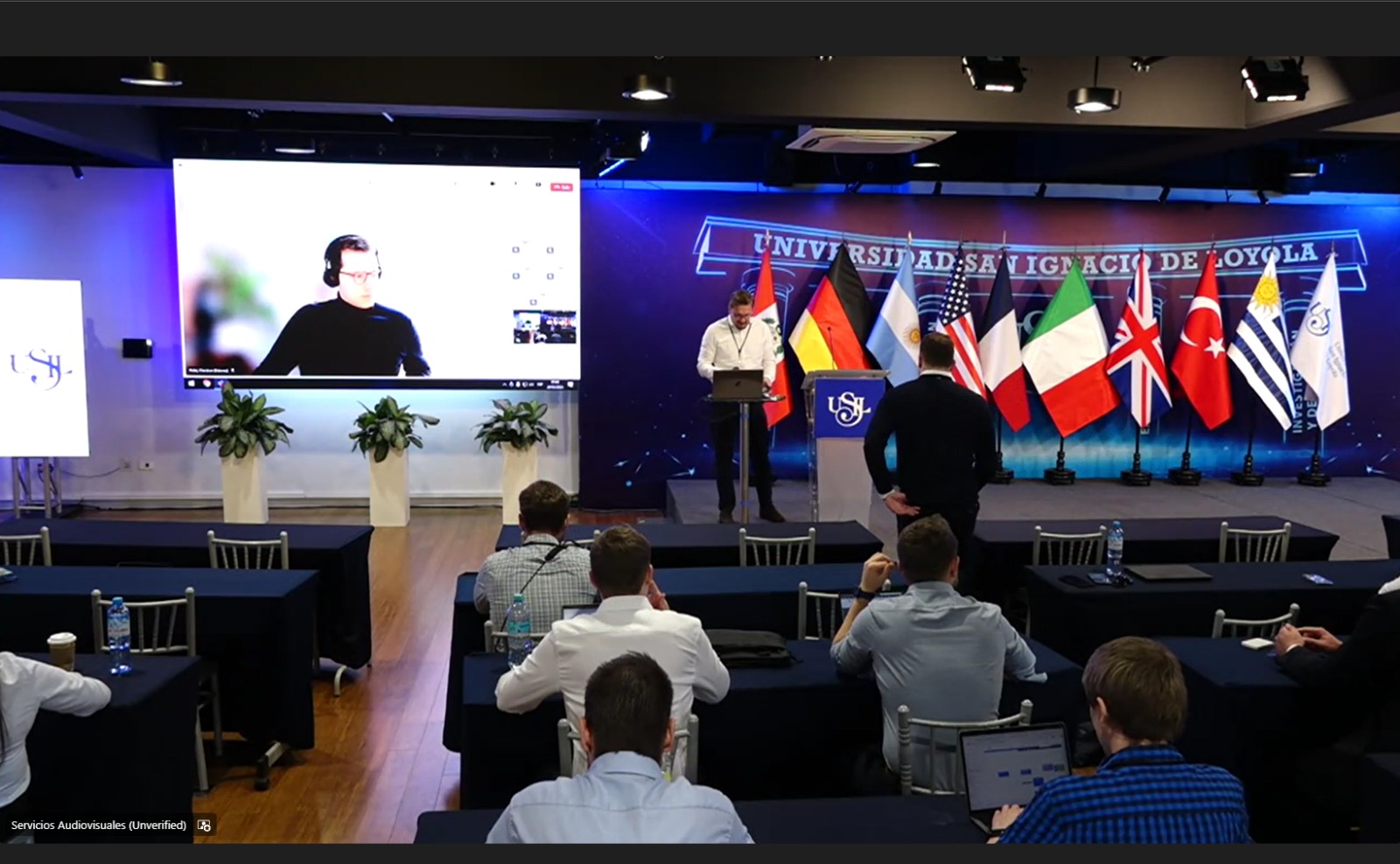 virtueller Konferenzvortrag, Christian Pulat ist auf einem Bildschirm zu sehen, im Plenum sitzen Personen, Flaggen mehrerer Nationen sind zu sehen