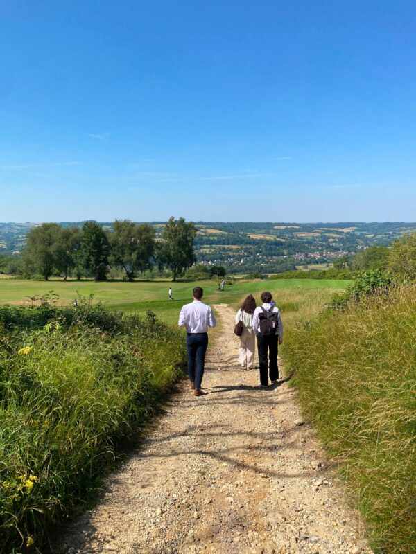 Eine Gruppe aus drei Menschen, zwei Männer, eine Frau, gehen einen Feldweg entlang, blauer Himmel, grüne Wiese, Gräser, Bäume