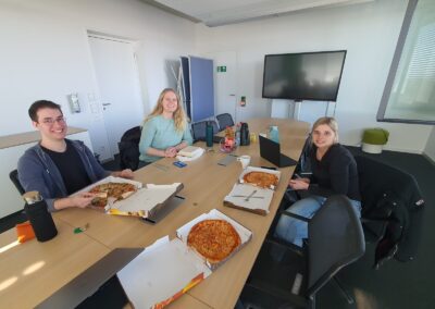 Personen sitzen um großen Besprechungstisch aus Holz, weiße Wände, es wurde Essen bestellt