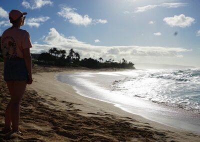 Fabienne Schnieders, trägt Cap, T-shirt und Shorts, steht am Strand von Maui, Sand, Wellen, Palmen, die Sonne scheint