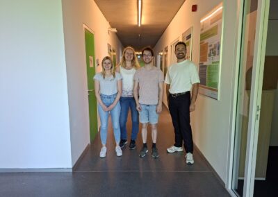 Fabienne Schnieders, Lara Kuhlmann (Frauen mit blonden Haaren), Jakob Becker und Leif Struwe stehen im Flur der Graduate School of Logistics, grüne Bürotüren